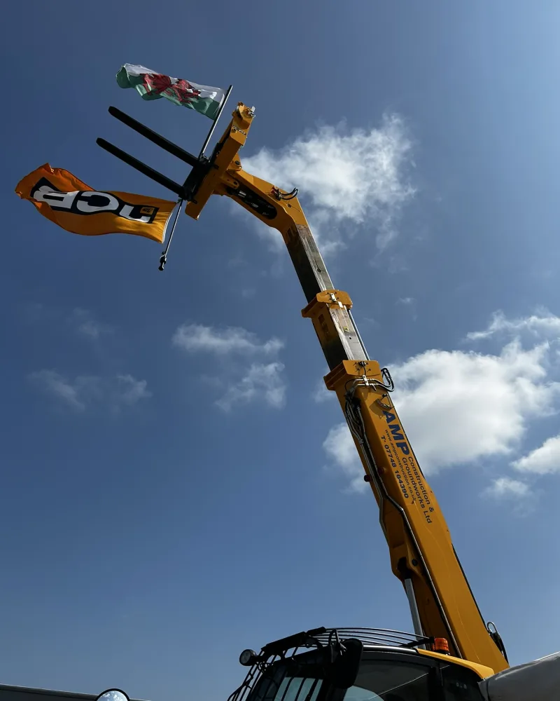 An AMP Construction telehandler with its boom raised, flying the Welsh flag, against a blue sky