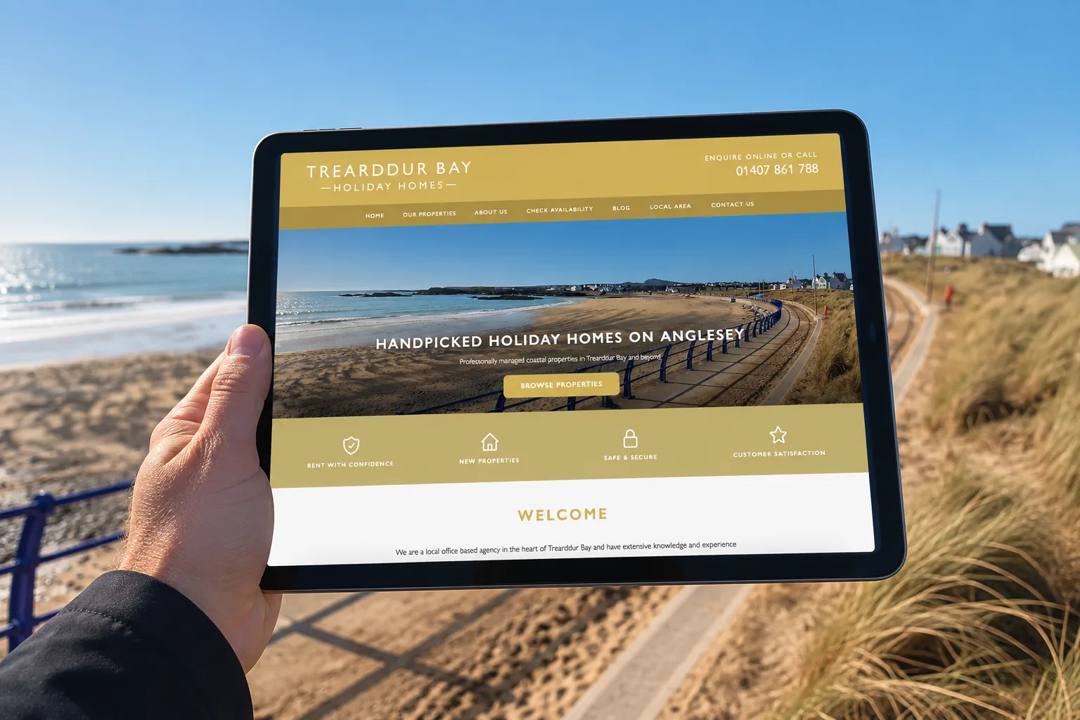 The Trearddur Bay Holiday Homes website on a tablet, held up on the promenade at Trearddur Bay